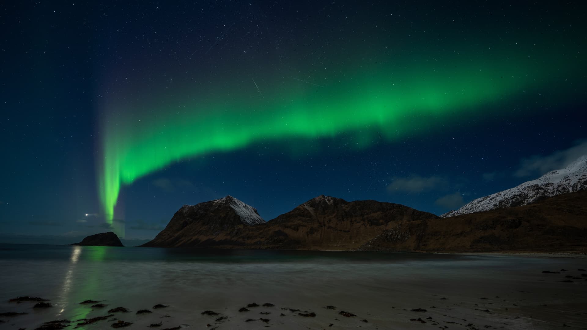 Green Northern Lights over a snow-covered coastline with mountains and a calm sea